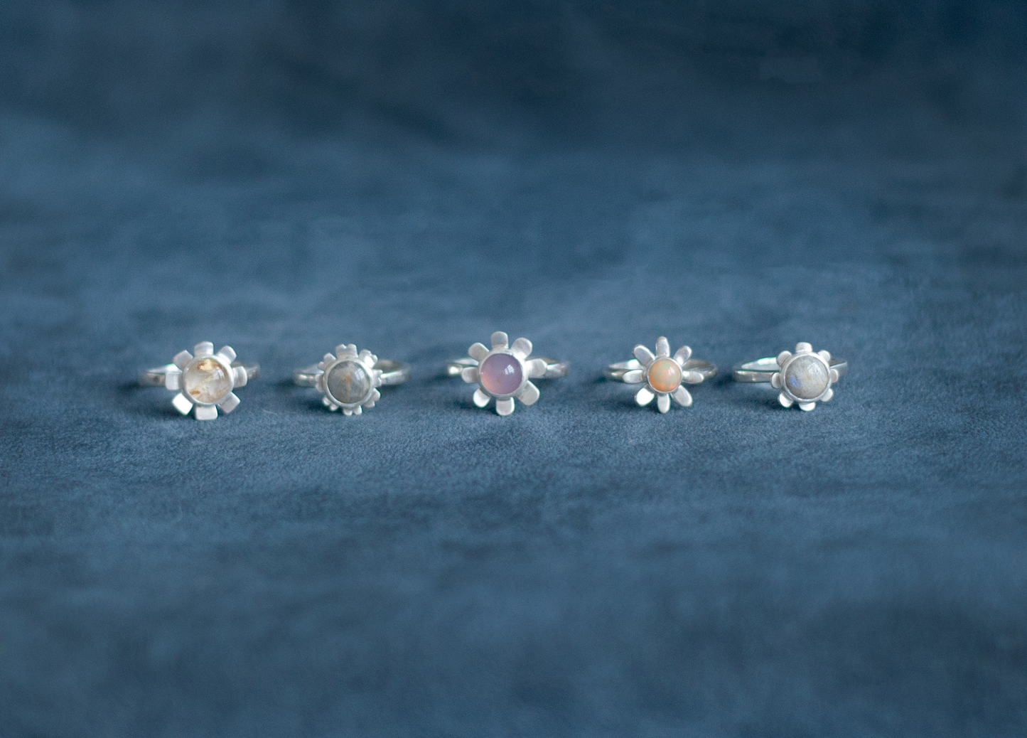 Five silver flower-shaped rings with different colored stones on a blue fabric background