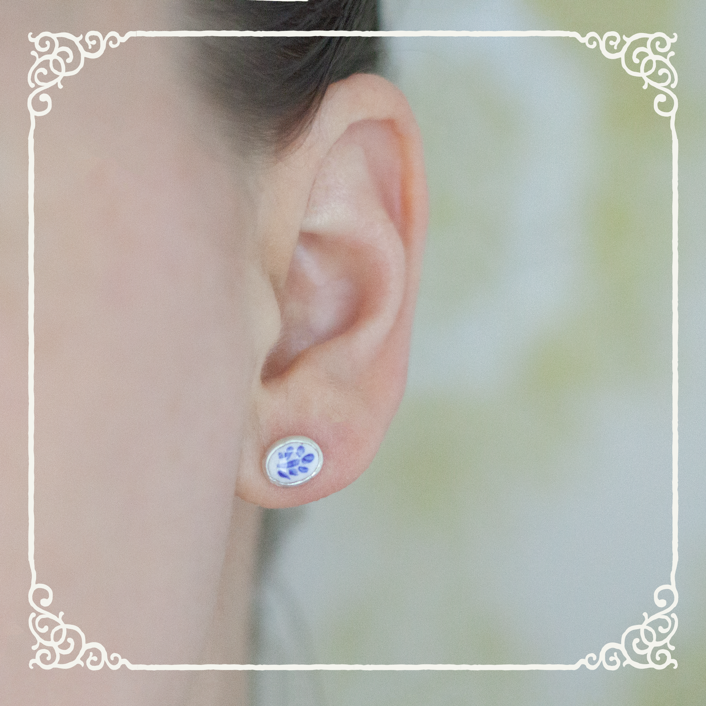 A close-up of a person's ear wearing a small oval stud earring with a blue floral pattern on a white background, framed by an ornate white border.